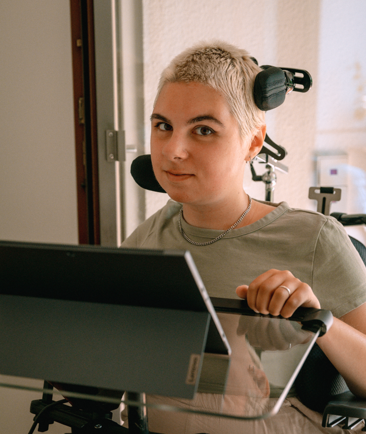 Eliza, eine weiße Person mit kurzen blonden Haaren sitzt im Rollstuhl, davor steht ein Rollstuhl mit Laptop drauf.